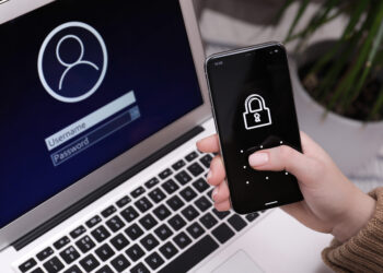 Dizüstü bilgisayarın giriş ekranı önünde, kilit simgesi görünen bir telefon tutan el.