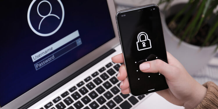 Dizüstü bilgisayarın giriş ekranı önünde, kilit simgesi görünen bir telefon tutan el.